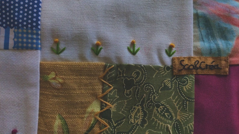 Pode incluir: Uma colcha de retalhos com uma variedade de tecidos, incluindo vichy azul e branco, flores marrons e brancas e paisley verde. A colcha &eacute; decorada com flores amarelas bordadas e caules verdes. A colcha tem uma etiqueta que diz "Scroll Creek".