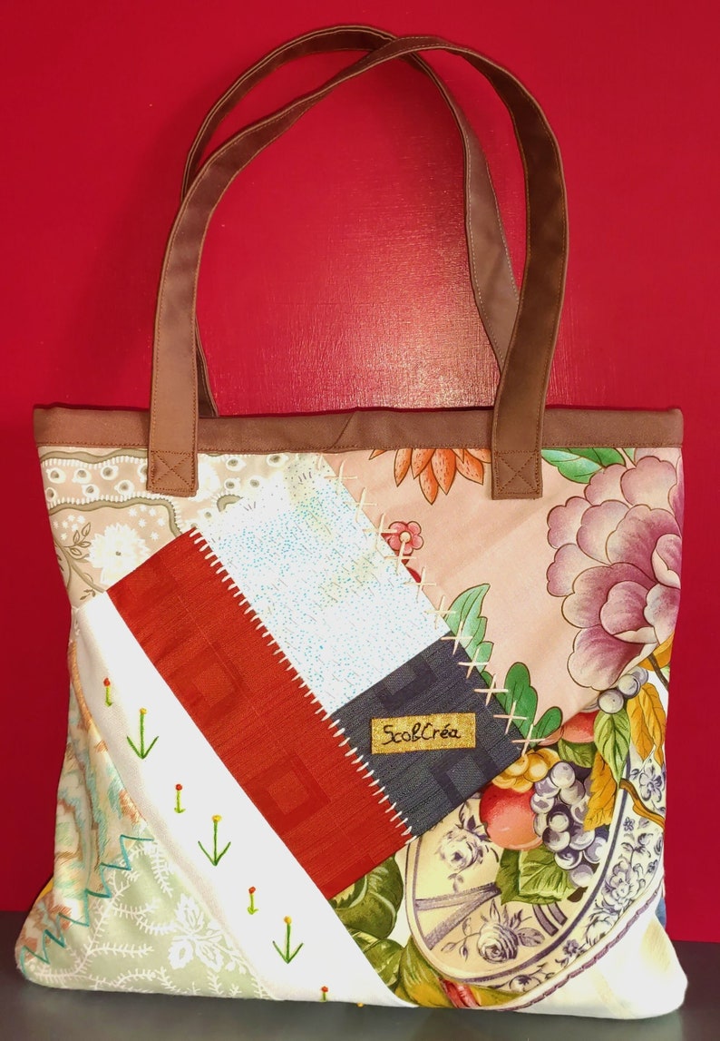 Pode incluir: Uma bolsa tote de patchwork com al&ccedil;as de couro marrom. A bolsa &eacute; feita de diferentes tecidos com padr&otilde;es florais, um tecido vermelho e branco e um tecido azul e branco. A bolsa tem uma etiqueta que diz "ScobCr&eacute;a".