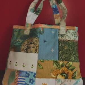 Pode incluir: Uma bolsa tote de patchwork com um design floral branco, azul e verde. A bolsa tem uma al&ccedil;a de lona branca e um design de patchwork com um sol, estrelas e flores.