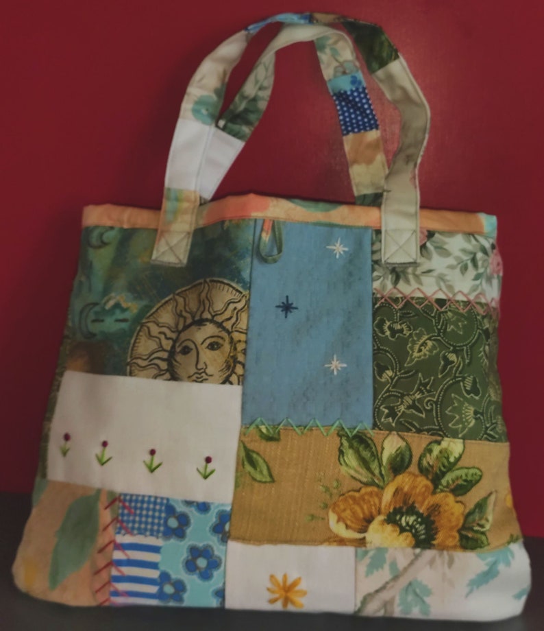 Pode incluir: Uma bolsa tote de patchwork com um design floral branco, azul e verde. A bolsa tem uma al&ccedil;a de lona branca e um design de patchwork com um sol, estrelas e flores.