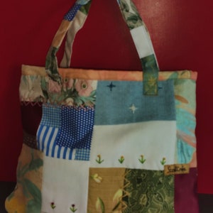 Pode incluir: Uma bolsa tote de patchwork com uma variedade de padr&otilde;es florais e geom&eacute;tricos em tons de azul, marrom, verde e branco. A bolsa tem uma al&ccedil;a de tecido branco e uma pequena etiqueta bordada que diz "Scotch Creek".