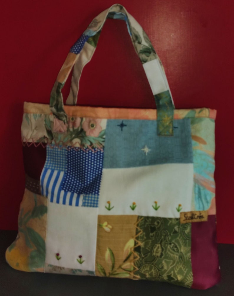 Pode incluir: Uma bolsa tote de patchwork com uma variedade de padr&otilde;es florais e geom&eacute;tricos em tons de azul, marrom, verde e branco. A bolsa tem uma al&ccedil;a de tecido branco e uma pequena etiqueta bordada que diz "Scotch Creek".