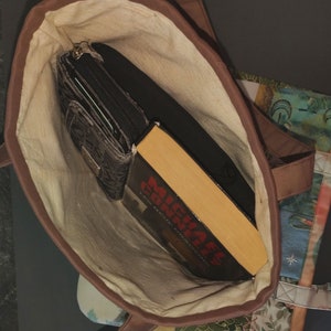 Pode incluir: Uma bolsa tote de lona marrom com forro de cor creme. A bolsa est&aacute; aberta e cont&eacute;m uma carteira preta e um livro de bolso intitulado "Michael Connelly The Lincoln Lawyer".