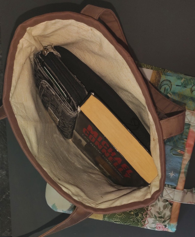 Pode incluir: Uma bolsa tote de lona marrom com forro de cor creme. A bolsa est&aacute; aberta e cont&eacute;m uma carteira preta e um livro de bolso intitulado "Michael Connelly The Lincoln Lawyer".
