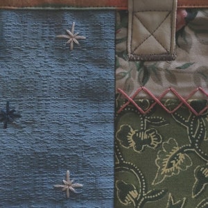 Pode incluir: Uma bolsa tote de tecido azul com um design de patchwork. A bolsa tem uma al&ccedil;a de couro marrom e um detalhe costurado rosa. O tecido &eacute; texturizado e tem um padr&atilde;o de estrelas.