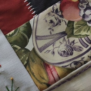 Pode incluir: Um cobertor de retalhos com uma variedade de tecidos, incluindo uma estampa floral com um prato e frutas, um tecido vermelho e um tecido branco com flores bordadas verdes.