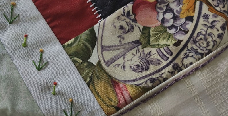 Pode incluir: Um cobertor de retalhos com uma variedade de tecidos, incluindo uma estampa floral com um prato e frutas, um tecido vermelho e um tecido branco com flores bordadas verdes.