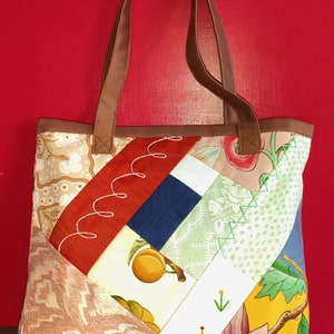 Pode incluir: Uma bolsa tote de patchwork com al&ccedil;as de couro marrom. A bolsa &eacute; feita de diferentes tecidos com padr&otilde;es florais e geom&eacute;tricos em tons de vermelho, azul, verde, amarelo e branco.