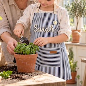 May include: A navy blue and white vertical striped apron featuring the name Sarah in white rope lettering across the chest, a small white flower embellishment, and a front pocket with a brown rectangular label, worn while gardening.