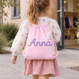 Può includere: Uno zaino con chiusura a coulisse rosa chiaro con il nome Anna ricamato in corsivo viola. La borsa presenta una piccola applicazione in peluche bianco a forma di coniglietto sulla parte anteriore ed è dotata di spessi cordini color crema.