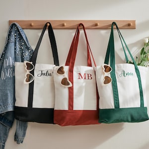 Op de afbeelding: Drie gepersonaliseerde draagtassen met witte canvas bodies en gekleurde accenten. Eén heeft zwarte afwerking en de naam "Julia", een andere heeft rode afwerking met "MB", en de derde heeft groene afwerking met "Anna". Elke tas heeft een decoratief houten accent.
