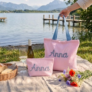 Könnte beinhalten: Eine Tragetasche und eine passende Reißverschlusstasche aus rosa-weißem Seersucker-Stoff, beide mit dem in blauer Schreibschrift gestickten Namen Anna versehen. Die Tragetasche verfügt über zwei blaue Stoffhenkel für ein stimmiges Design.