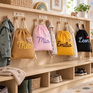 Pode incluir: Quatro mochilas de cordão nas cores mostarda, rosa, amarelo e preto, personalizadas com os nomes bordados Theo, Mia, Emilia e Leon. As bolsas apresentam pequenas aplicações de pelúcia de urso, coelho, flor e carro, penduradas em ganchos de madeira.