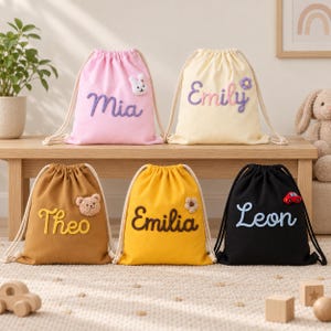 Könnte beinhalten: Fünf Kordelzugrucksäcke in Rosa, Creme, Braun, Gelb und Schwarz. Jede Tasche zeigt einen in Schreibschrift gestickten Namen: Mia, Emily, Theo, Emilia und Leon. Kleine Plüschapplikationen in Form von Hase, Blume, Bär, Blume und Auto sind angebracht.