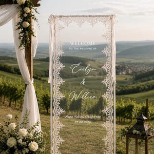 May include: A rectangular clear acrylic wedding welcome sign framed in white with intricate white lace trim along the edges. The text reads: WELCOME TO THE WEDDING OF Evelyn & William, Napa Valley, California, 02.08.2026, printed in white.