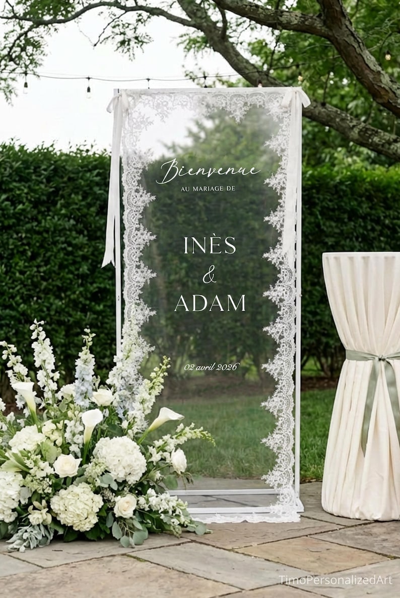 May include: A rectangular clear acrylic wedding welcome sign framed in white with white lace trim along the vertical edges. The sign features the text Bienvenue AU MARIAGE DE IN&Egrave;S & ADAM 02 avril 2026 in white lettering with white ribbon ties.