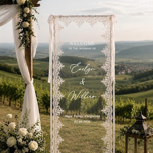 May include: A rectangular clear acrylic wedding welcome sign framed in white with intricate white lace trim along the edges. The text reads: WELCOME TO THE WEDDING OF Evelyn & William, Napa Valley, California, 02.08.2026, printed in white.