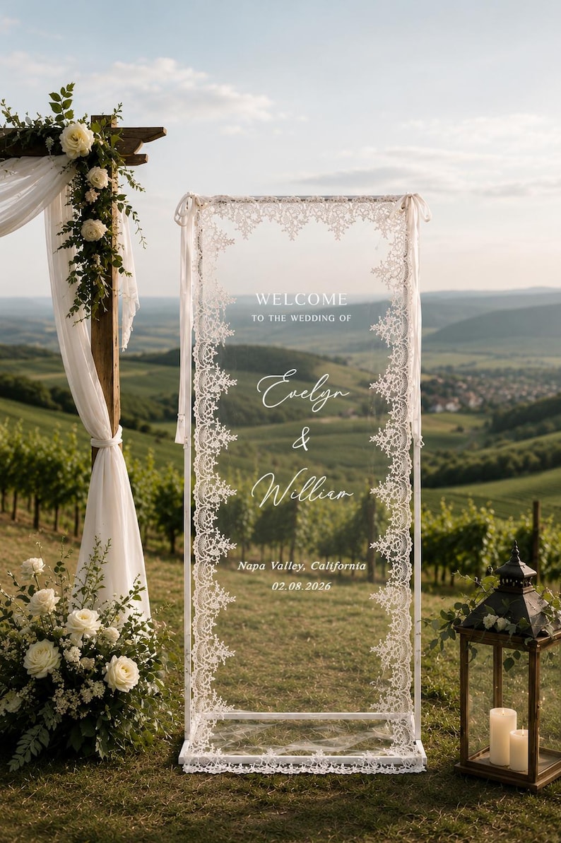 May include: A rectangular clear acrylic wedding welcome sign framed in white with intricate white lace trim along the edges. The text reads: WELCOME TO THE WEDDING OF Evelyn & William, Napa Valley, California, 02.08.2026, printed in white.