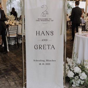 Può includere: Un alto cartello di benvenuto per matrimoni in tessuto color bianco sporco, rettangolare, sospeso a una struttura metallica con nastri verde salvia. Il testo recita: Welcome to the wedding of Hans and Greta, Schwabing, München, 18. 10. 2025, con un'icona floreale.