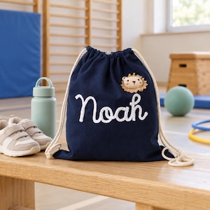 Könnte beinhalten: Ein dunkelblauer Turnbeutel mit dem Namen Noah in weißer Schreibschrift und einem kleinen Plüsch-Löwenkopf-Aufnäher mit Krone. Der Beutel verfügt über dicke, cremefarbene Kordelzüge und besteht aus einem festen Stoff mit einem gerafften Verschluss.