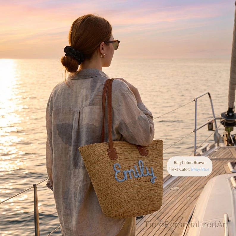 May include: A woven straw tote bag with brown leather-like handles and the name Emily embroidered in baby blue script lettering on the front. The bag features a textured natural tan straw construction and two vertical brown shoulder straps.