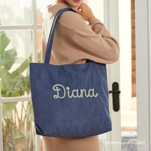 May include: A blue corduroy tote bag with the name "Diana" embroidered in a light green script. The bag has long handles and is being held by a person. The bag is a medium size and appears to be made of a soft material.