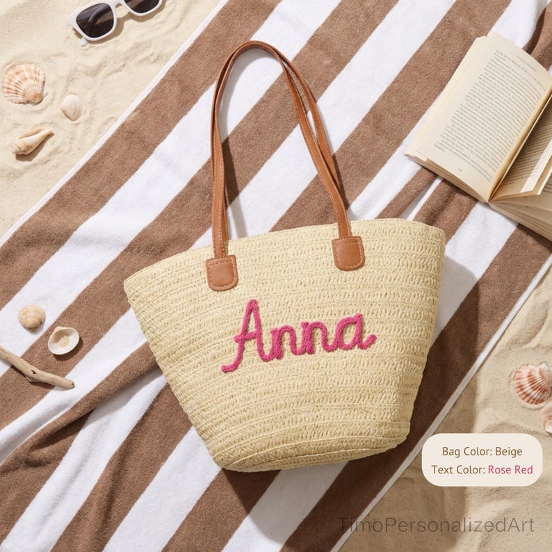 May include: A beige woven straw tote bag featuring two brown handles and the name Anna embroidered in rose red script lettering on the front. The bag has a structured base and a textured surface, showcasing a natural fiber construction.