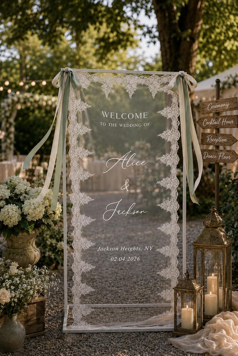 May include: A rectangular clear acrylic wedding welcome sign in a white metal frame, decorated with white lace trim along the vertical edges and sage green ribbons at the top corners. The text reads: WELCOME TO THE WEDDING OF Alice & Jackson, Jackson Heights, NY, 02.04.2026.
