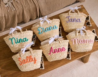 May include: A collection of woven straw tote bags, each with a different name embroidered in colorful thread. The bags have white ribbon handles and are arranged on a wooden surface. The names include "Nina", "Diana", "Jana", "Anne", "Mama", and "Sally".