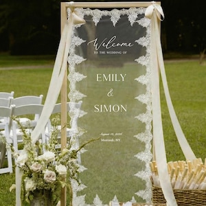 May include: A rectangular clear acrylic wedding welcome sign framed in light wood, featuring white lace trim along the edges and cream ribbons. The text reads: Welcome TO THE WEDDING OF EMILY & SIMON August 12, 2026 Montauk, NY.