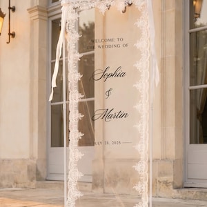 May include: A tall, rectangular, transparent acrylic wedding welcome sign framed by white lace trim. The sign features black text reading "WELCOME TO THE WEDDING OF Sophia & Martin JULY 28, 2025" and is supported by a white metal stand with white ribbons.