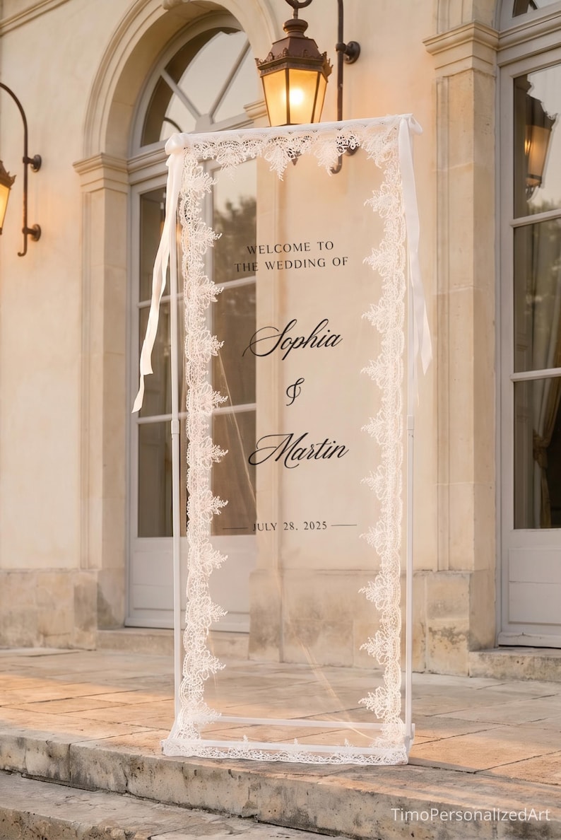 May include: A tall, rectangular, transparent acrylic wedding welcome sign framed by white lace trim. The sign features black text reading "WELCOME TO THE WEDDING OF Sophia & Martin JULY 28, 2025" and is supported by a white metal stand with white ribbons.