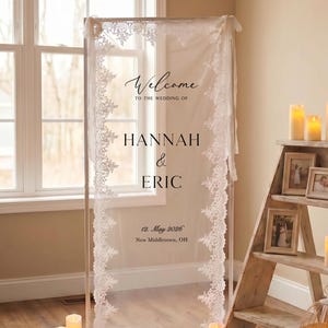 May include: A rectangular wedding welcome sign featuring a sheer white fabric panel framed in a thin metallic structure. The fabric is bordered by intricate white lace trim and displays the text Welcome to the wedding of Hannah & Eric 12. May 2026.