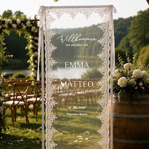 May include: A rectangular white frame holding a sheer fabric panel with intricate white lace borders. The text reads Willkommen ZUR HOCHZEIT VON EMMA & MATTEO 12. Mai 2026 Tegernsee, Bayern. The frame is supported by a base and tied with white ribbons.