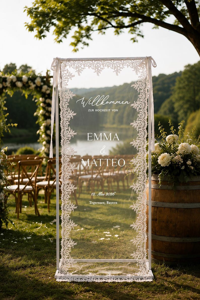 May include: A rectangular white frame holding a sheer fabric panel with intricate white lace borders. The text reads Willkommen ZUR HOCHZEIT VON EMMA & MATTEO 12. Mai 2026 Tegernsee, Bayern. The frame is supported by a base and tied with white ribbons.