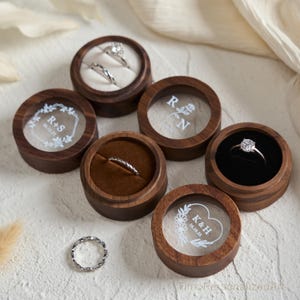 Könnte beinhalten: Eine Sammlung runder Ringkästchen aus Holz mit transparenten Deckeln, die weiße Gravuren wie die Initialen R+S, R N und K&H mit Daten zeigen. Einige Kästchen enthalten silberne Ringe mit Steinen, andere haben weiße oder schwarze Samteinlagen.