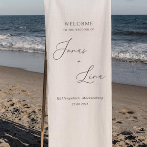 Peut inclure: Une bannière de mariage verticale en tissu blanc cassé suspendue à une tige en bois. Le texte indique : WELCOME TO THE WEDDING OF Jonas + Lina, Kühlungsborn, Mecklenburg, 22.08.2025. La bannière est fixée par des rubans blancs aux coins supérieurs.