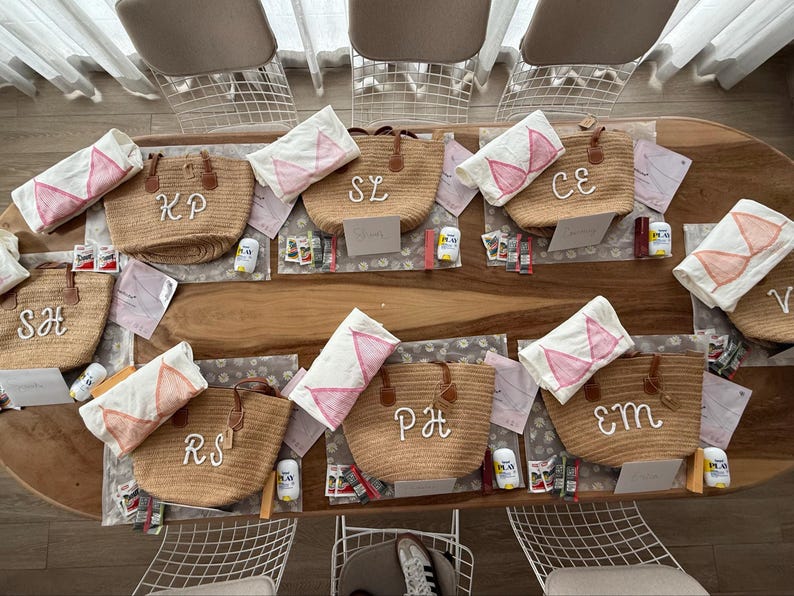 May include: A collection of woven straw tote bags with brown leather handles, each featuring white embroidered initials. Accompanying each bag is a white fabric pouch with a pink bikini top graphic, along with small personal care items.