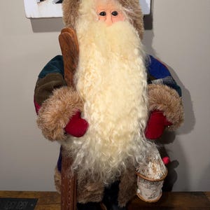 Peut inclure: Figurine du Père Noël en peluche avec une longue barbe crème et un chapeau et des mitaines bordés de fourrure brune. Il porte un manteau à carreaux et des bottes noires, tenant des skis en bois et une lanterne en écorce de bouleau. Décoration de vacances festive.