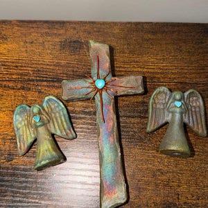 May include: A set of three religious decorative items: a cross and two angel figurines. The cross is made of a textured, iridescent material with a turquoise center and copper wire accents. The angels have turquoise hearts.