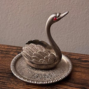 May include: A silver swan figurine with a red gem eye, perched on a decorative round tray. The swan has detailed feather carvings and a curved neck. The tray has an ornate, embossed pattern.