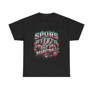 May include: Black t-shirt featuring a colorful graphic design. The design includes the text "SPURS BASKETBALL" with a cityscape and basketball elements. The Spurs logo is prominently displayed in the center.