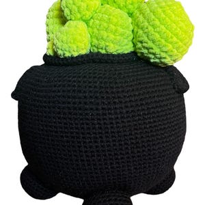 May include: A black crocheted cauldron with a green crocheted plant inside. The cauldron has three legs and a rounded bottom.