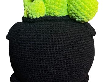 May include: A black crocheted cauldron with a green crocheted plant inside. The cauldron has three legs and a rounded bottom.