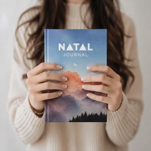 May include: A hardcover journal with the title "NATAL JOURNAL" in white letters. The cover features a landscape painting of a blue sky, clouds, and a forest silhouette. The journal is held by a person wearing a cream-colored sweater.