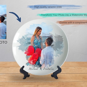 Könnte beinhalten: Ein weißer Keramikteller mit einem Aquarellbild eines Paares, das rote Ballons an einem Strand hält. Der Teller trägt die Namen "Michael & Isabella" und wird mit einem Ständer geliefert.