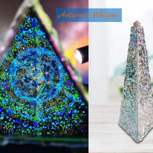 Può includere: Una piramide di vetro con un design olografico che presenta un motivo a mandala blu e verde. La piramide è su una superficie di legno. Il testo "Autumn's Whisper" è visibile sullo sfondo.