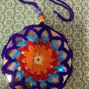 May include: A handmade, circular ornament featuring a repurposed CD, crocheted with vibrant purple yarn. The center showcases a colorful crochet design with orange, red, and blue accents. A purple cord with rainbow beads is attached for hanging.