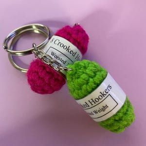May include: A green and pink crocheted keychain with a label that reads "Crooked Hookers Worsted Weight".