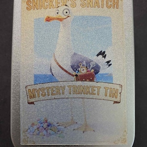Puede incluir: Una lata rectangular plateada con una ilustración de una gaviota de dibujos animados. La gaviota lleva un cofre del tesoro. El texto en la lata dice "SNICKER'S SNATCH" y "MYSTERY TRINKET TIN" en un estilo vintage.
